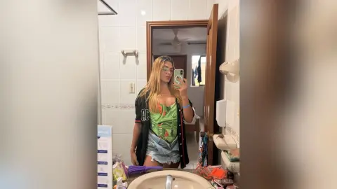 Young woman in a green floral top and denim shorts taking a mirror selfie in a bright bathroom.