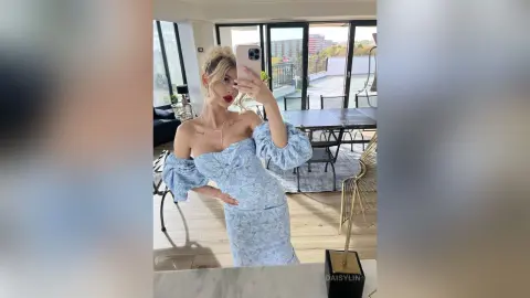 Blonde woman in a light blue floral off-the-shoulder dress taking a selfie in a bright modern living room.