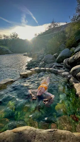 Woman in an orange bikini relaxing in a natural rock pool under bright sunlight in a lush canyon setting.