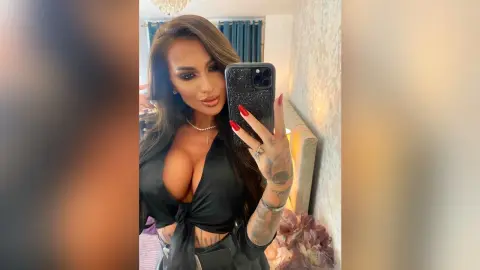 Glamorous brunette woman in a black low-cut blouse taking a confident mirror selfie in a bright room.