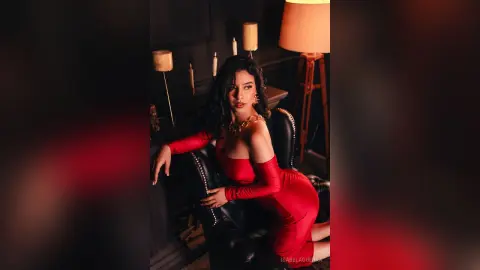 Elegant woman in a red off-the-shoulder dress sitting in a moody, dimly lit room with warm candlelight.