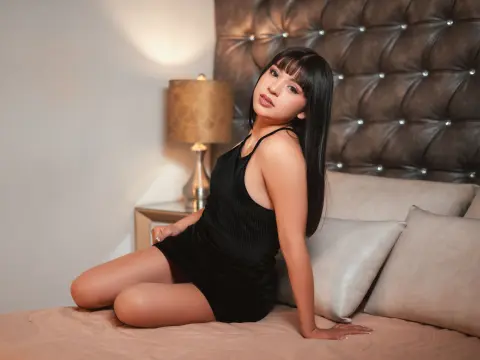 VelvettGal poses on a bed wearing a short black tank top, showing off her long dark hair and smooth legs against a tufted headboard.