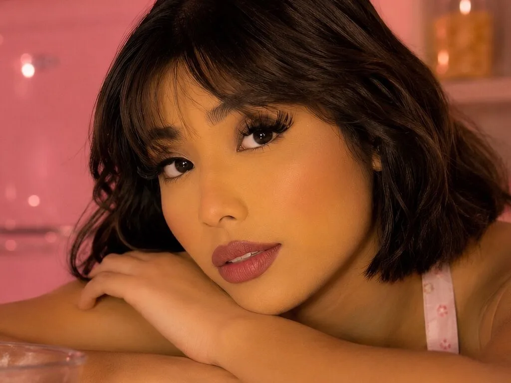 CorinaBaker — CorinaBaker rests her chin on her hand, looking directly at the camera with dark wavy hair and pink lipstick.