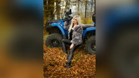 Blonde woman in a grey faux fur coat and black leather pants sitting on a blue ATV in an autumn forest.
