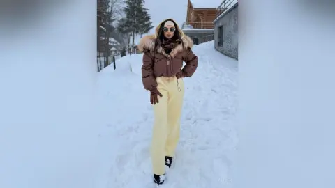 Stylish woman in a brown puffer jacket and cream trousers walking through a snowy winter landscape.