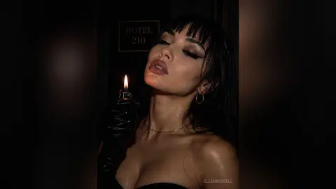 Glamorous woman in black gloves holding a lit lighter in a moody, dark hotel hallway setting.