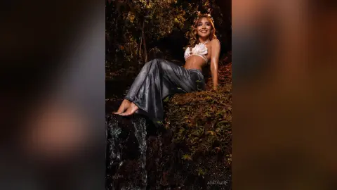 AdaTylor sits naked on a mossy rock ledge near a small waterfall, wearing a white lace bra and grey silk pants.