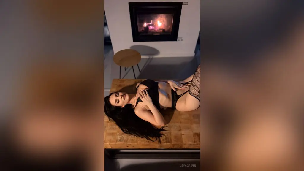 Leya — Woman in black lingerie relaxing on a wooden table near a cozy glowing fireplace in a dim room.