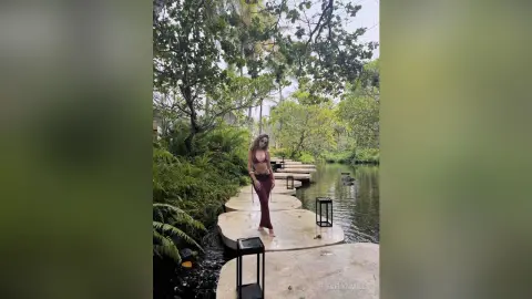 Woman in a burgundy bikini standing on a stone path by a tropical jungle pond during a serene summer day.