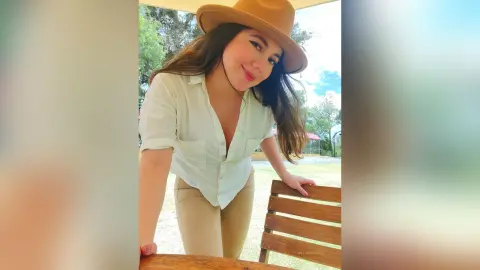 Smiling woman in a white button-down shirt and tan trousers wearing a wide-brimmed hat in a sunny outdoor park.