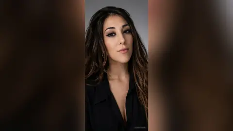 Portrait of a brunette woman in a black shirt with dramatic eyeliner against a neutral studio background.