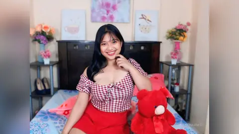 Smiling young woman in a red gingham top sitting on a bed with a red teddy bear in a cozy bedroom setting.