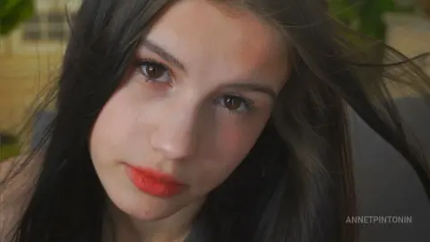 Close-up portrait of a young woman with long dark hair and red lipstick in soft natural lighting.
