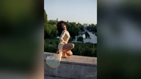 Curvy woman in a sheer white lace maxi dress sitting on a stone ledge in a lush green park at sunset.
