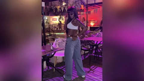 Stylish woman in a white crop top and baggy jeans posing in a moody, neon-lit outdoor lounge setting.