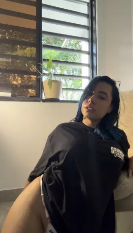 Young woman with blue hair wearing an oversized black t-shirt relaxing in a sunlit room by a window.