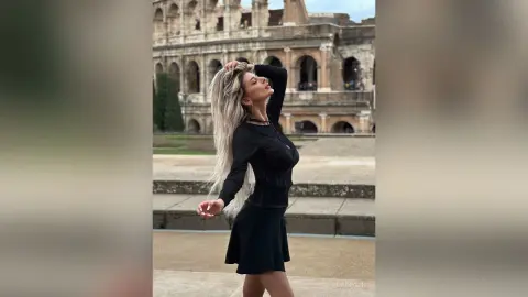 DaisyLin poses outdoors in front of the Colosseum wearing a long-sleeved black top and a short black skirt, running her hands through her long blonde hair.