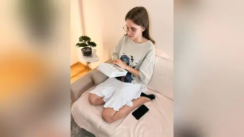 Young woman in a casual graphic tee and white skirt reading a book on a cozy sofa in a bright room.