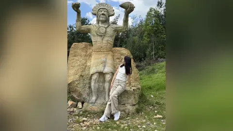 Woman in white tank top and khaki pants leaning against a large ancient stone statue in a lush green forest.