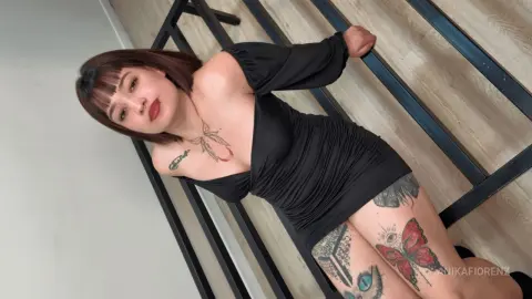 Tattooed woman in a black off-the-shoulder mini dress posing in a modern minimalist room.