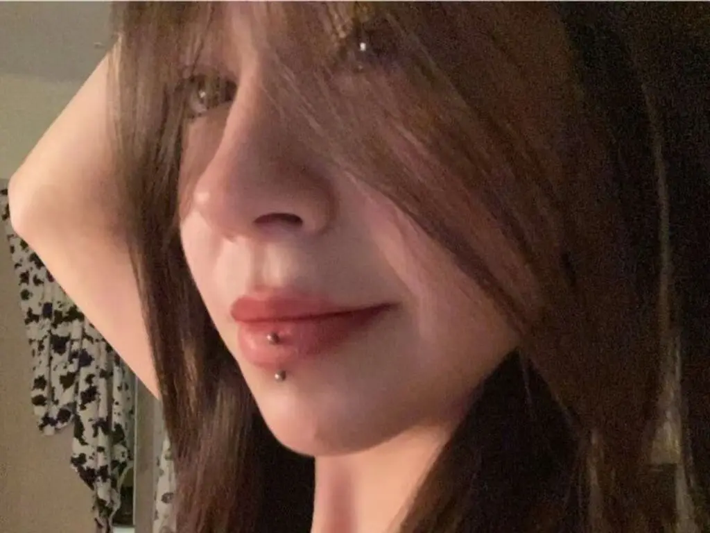 Close-up portrait of a woman with long brown hair and a lip piercing in soft indoor lighting.
