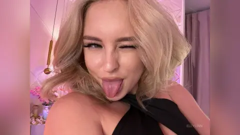 Blonde woman in a black halter top winking and sticking her tongue out in a bright pink room.