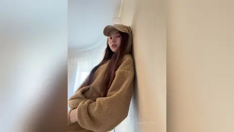Young woman in a cozy tan fleece sweater and baseball cap leaning against a wall in a bright room.