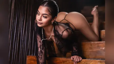 Elegant woman in black lace lingerie posing on wooden stairs in a moody, dimly lit indoor setting.