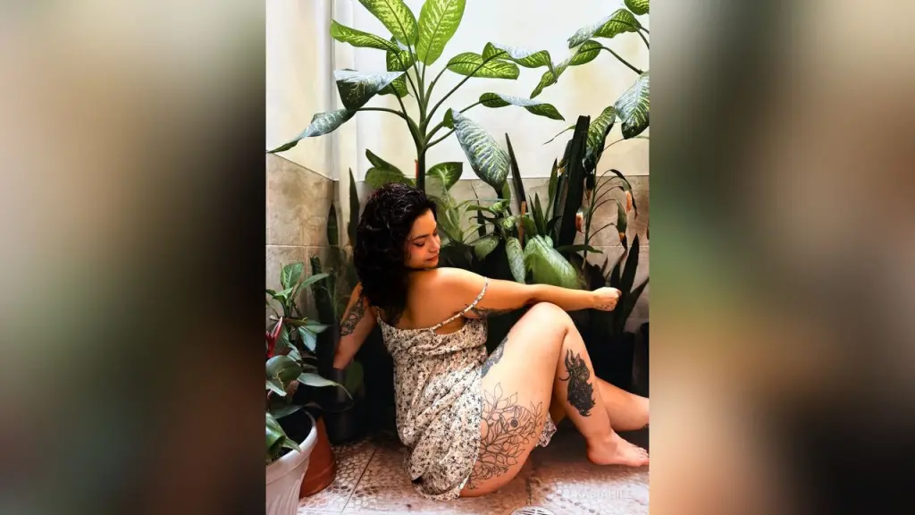 KasiaHill — Tattooed woman in a floral sundress sitting amidst lush green houseplants in a bright indoor setting.