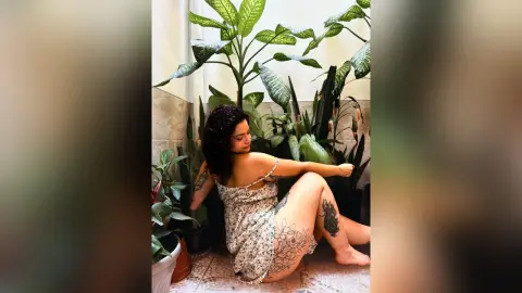 Tattooed woman in a floral sundress sitting amidst lush green houseplants in a bright indoor setting.