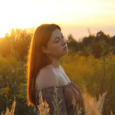 Young woman in an off-the-shoulder top gazing thoughtfully during a golden hour sunset in a wildflower field.