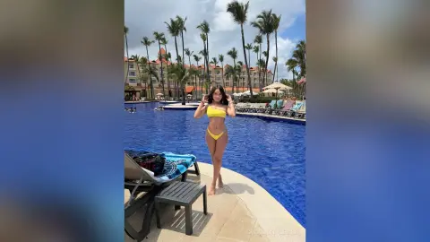 Young woman in a yellow bikini relaxing by a tropical resort swimming pool under palm trees.