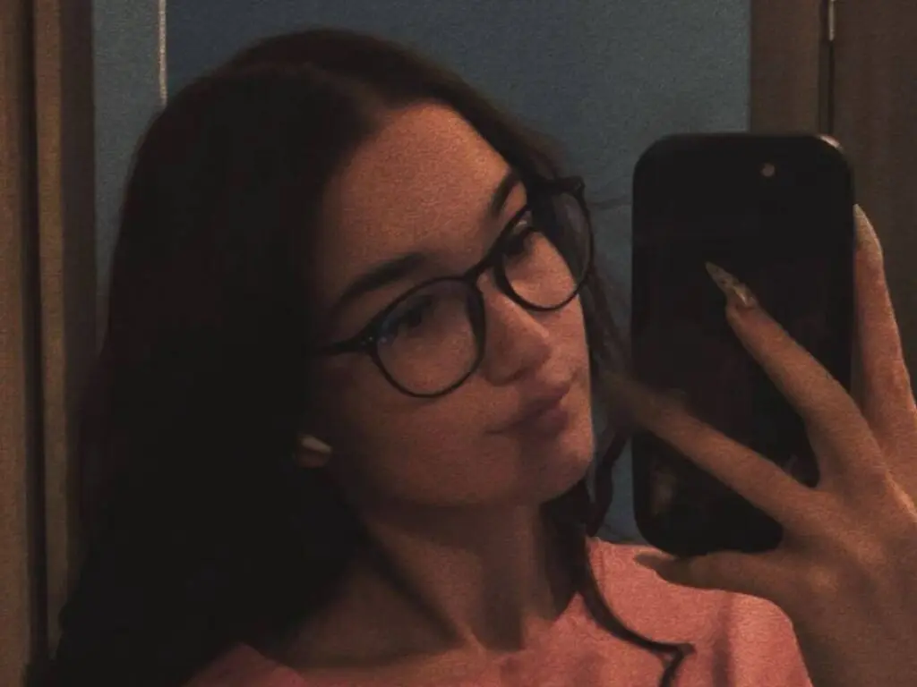 Young woman with glasses taking a moody selfie in a pink top indoors.