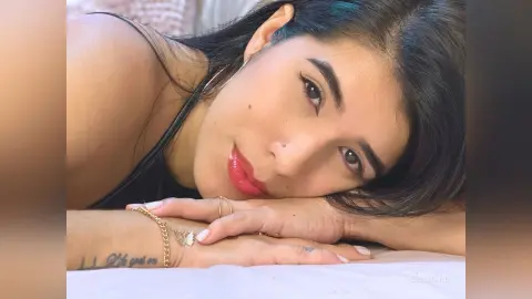 Smiling young woman with dark hair wearing a black tank top while relaxing on a white bed surface.