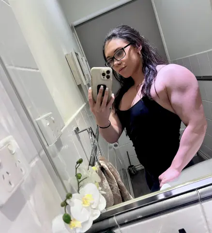 Fit woman in black tank top taking a mirror selfie in a bright bathroom.