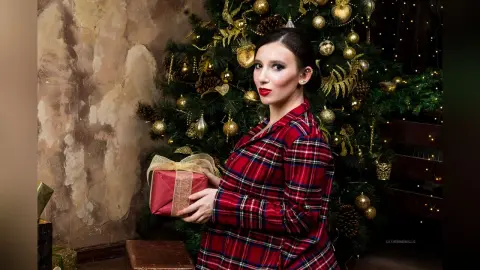 Elegant woman in red plaid pajama holding a gift near a glowing Christmas tree in a cozy festive setting.