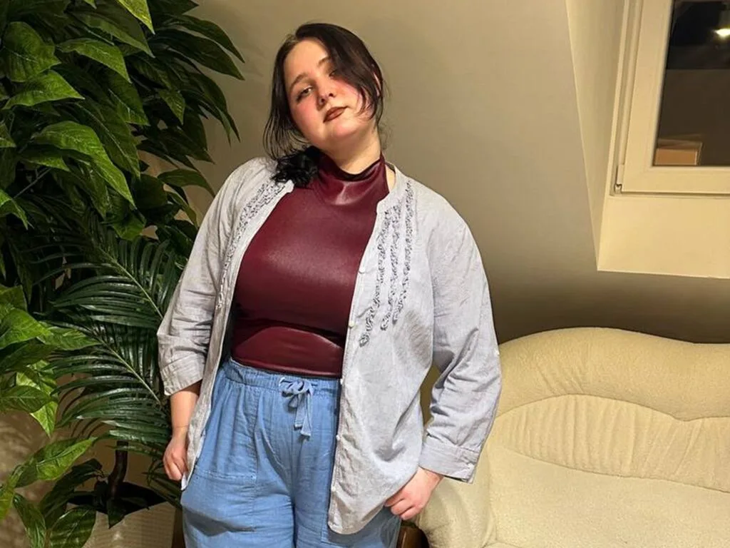 WildObsessionn wears a maroon mock-neck top and light blue linen trousers while standing next to a large green plant and a white sofa.