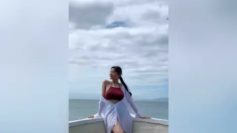 Young woman in a white linen shirt and burgundy bikini top relaxing by the ocean under a cloudy sky.