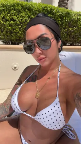 Tanned woman in white polka dot bikini and sunglasses relaxing on a sunny summer day by the pool.