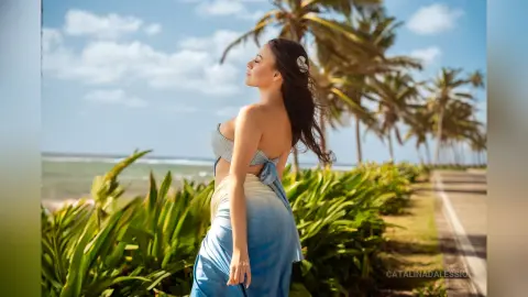 Dreamy portrait of a woman in a blue wrap skirt standing by tropical palm trees on a sunny beach.