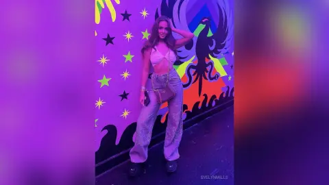 EvelynMills stands against a colorful graphic wall wearing a white lace bra and shimmering silver glittery trousers.