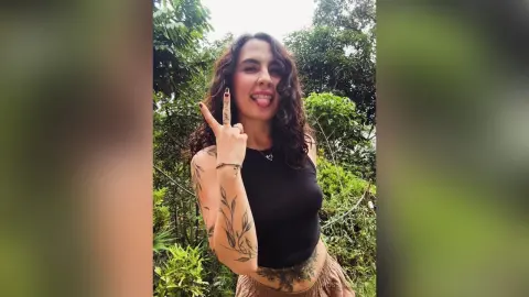 RossiNoleen sticks out her tongue while wearing a black tank top and posing against a background of green trees and leaves.