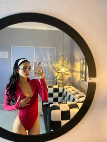 Young woman in a pink bodysuit and glasses taking a mirror selfie in a modern living room.