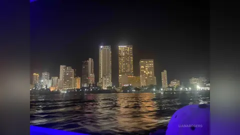 Nighttime cityscape view of illuminated skyscrapers reflecting on dark river water under a clear night sky.