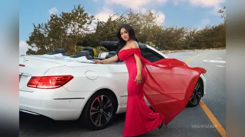 Miah stands next to a white convertible car wearing a long, off-the-shoulder red dress against a background of trees and a blue sky.
