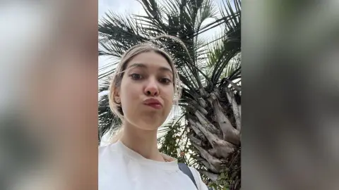 Young woman in a white t-shirt making a playful expression outdoors near palm trees.