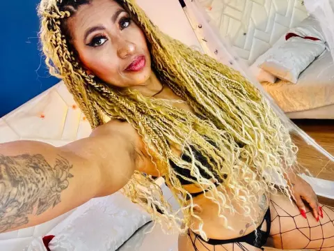 Woman with long blonde braids in black lingerie relaxing on a white quilted bed in a bright room.