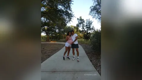 Happy pregnant woman in a pink top and man in a white t-shirt walking on a park path under leafy trees.