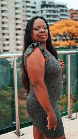 Smiling Black woman in a grey sparkly cocktail dress posing on a city balcony at sunset.