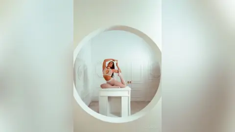 ElizaLuxe poses sitting on a white pedestal in a white room, wearing a white bra and matching undies with her hands behind her head.